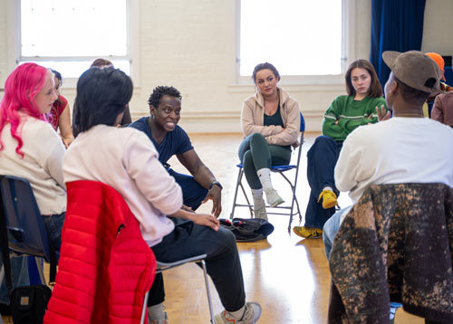 Actor Kobna Holdbrook-Smith kneeling in the middle of a circle of Actor students during a workshop.